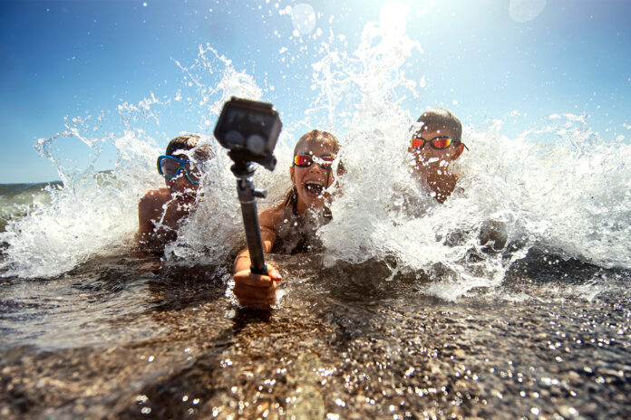 Trois jeunes en maillot de bain immortalisent un moment de plaisir avec une caméra portative, illustrant les incontournables techno pour un été exceptionnel alors que les vagues les entourent sur une plage ensoleillée.