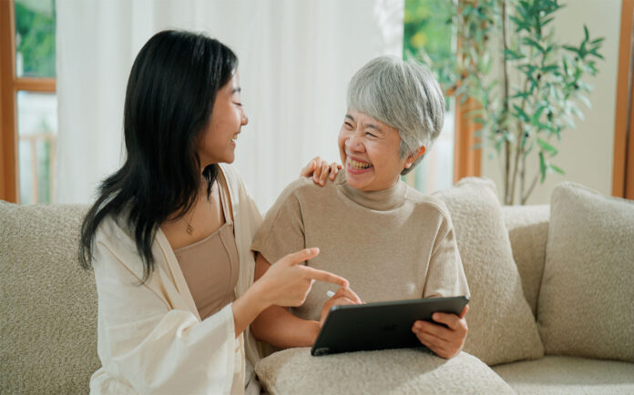 A young Asian woman teaching her retired mother how to use a digital tablet in the living room.