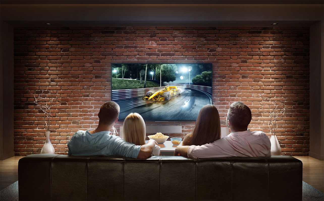 Two couples are cheering while watching game at home. They are sitting on a sofa in the modern living room faced to a big TV set on the front wall. The car is artificial and made in 3D.