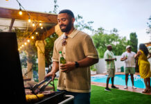 Man grilling food while holding a beer at an outdoor barbecue party by the pool. Group of friends socializing in the background. Summer celebration concept.
