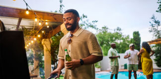 Man grilling food while holding a beer at an outdoor barbecue party by the pool. Group of friends socializing in the background. Summer celebration concept.