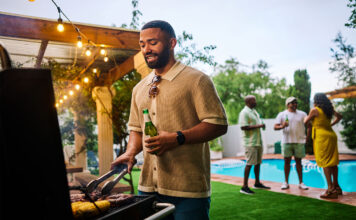 Man grilling food while holding a beer at an outdoor barbecue party by the pool. Group of friends socializing in the background. Summer celebration concept.