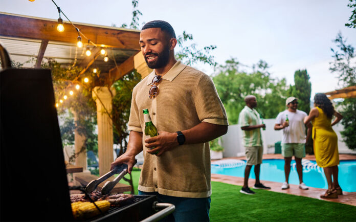Man grilling food while holding a beer at an outdoor barbecue party by the pool. Group of friends socializing in the background. Summer celebration concept.