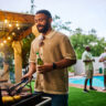 Man grilling food while holding a beer at an outdoor barbecue party by the pool. Group of friends socializing in the background. Summer celebration concept.