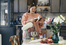 woman standing in front of a table in her kitchen, stand mixer on it. She is reading a recipe book in order to check a recipe for a dessert she is going to make.