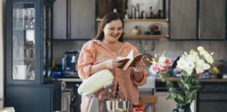woman standing in front of a table in her kitchen, stand mixer on it. She is reading a recipe book in order to check a recipe for a dessert she is going to make.