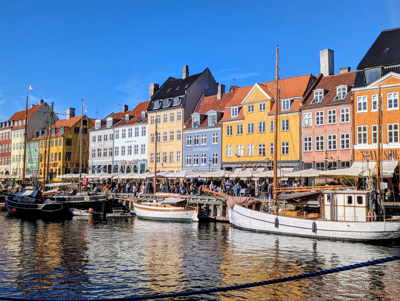 Photo of a canal in Copenhagen, with colorful houses, moored boats, and people walking along the water's edge.