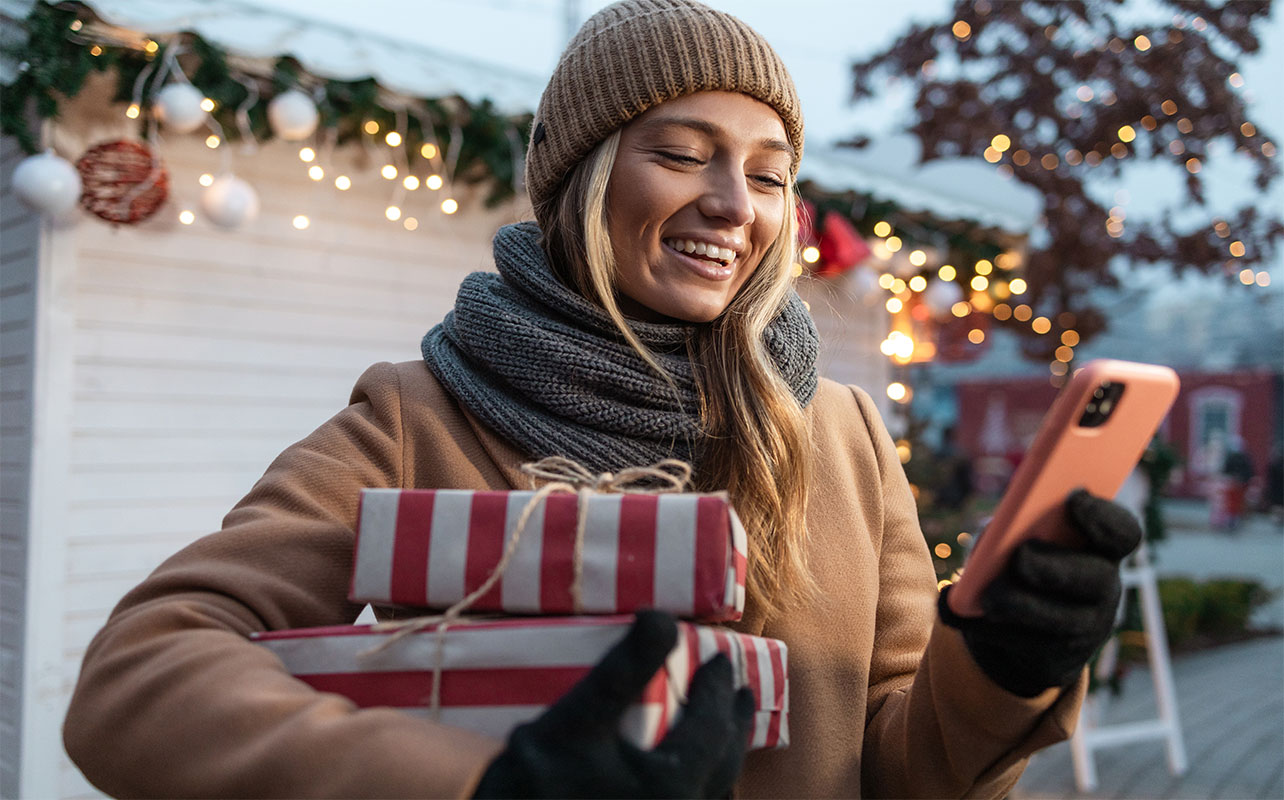 Photo d’une jeune femme heureuse tenant des cadeaux techno et regardant son téléphone, dans un décor des Fêtes en plein air