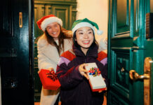 15 cadeaux de dernière minute pour faire vraiment plaisir Girlfriends in festive hats arriving at the door, holding wrapped gifts and holiday drinks.