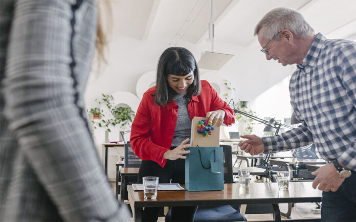 15 cadeaux de dernière minute pour faire vraiment plaisir A woman receiving last-minute gifts in a bag from her colleagues and celebrating at the office after a recent success.
